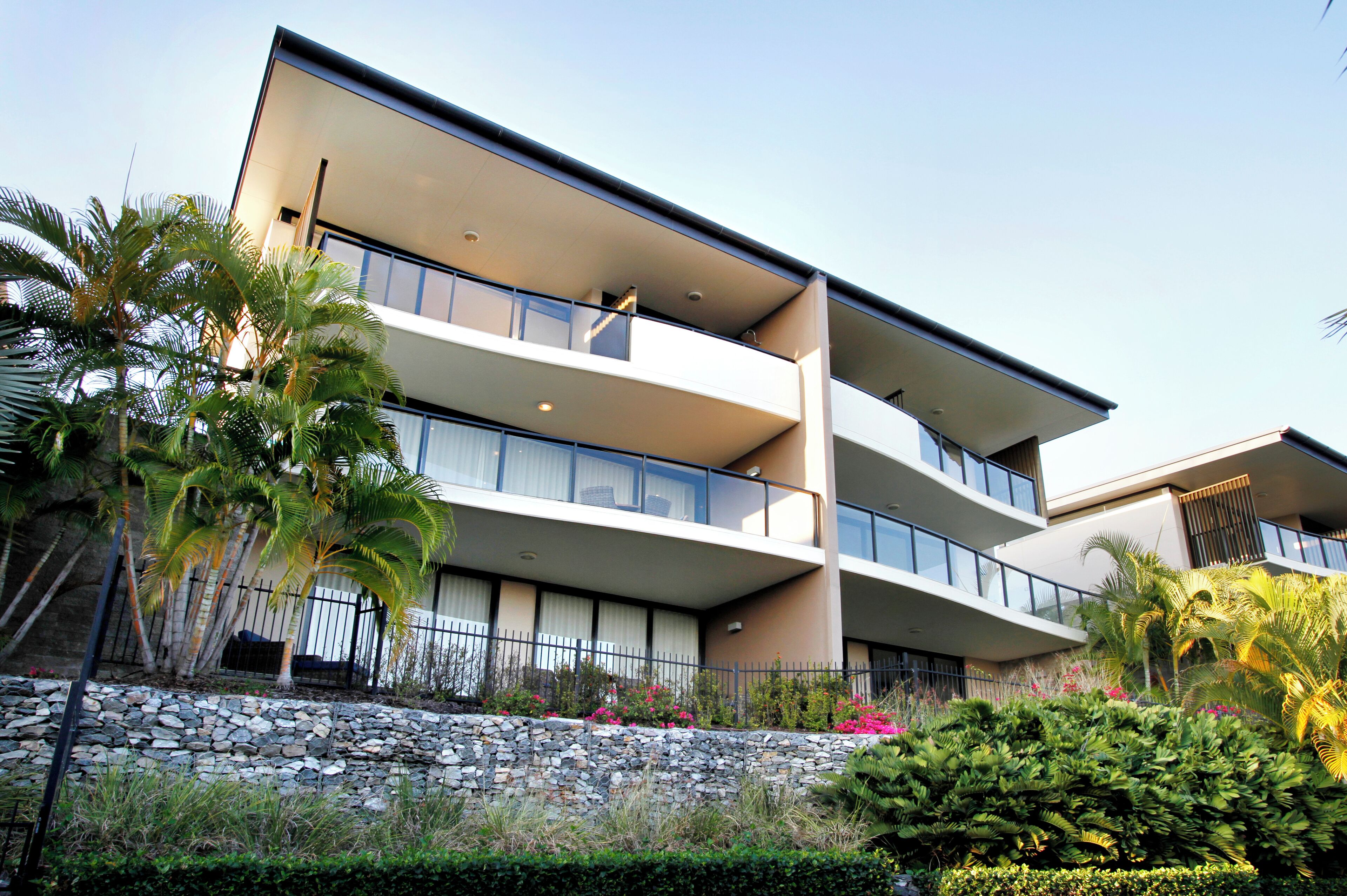 Un edificio de varios pisos con balcones, un muro de contención de piedra y vegetación tropical.