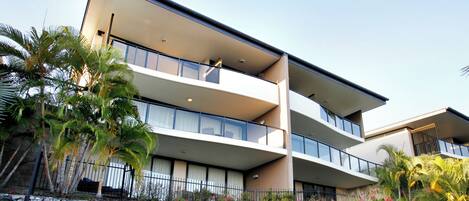 Un edificio de varios pisos con balcones, un muro de contención de piedra y vegetación tropical.