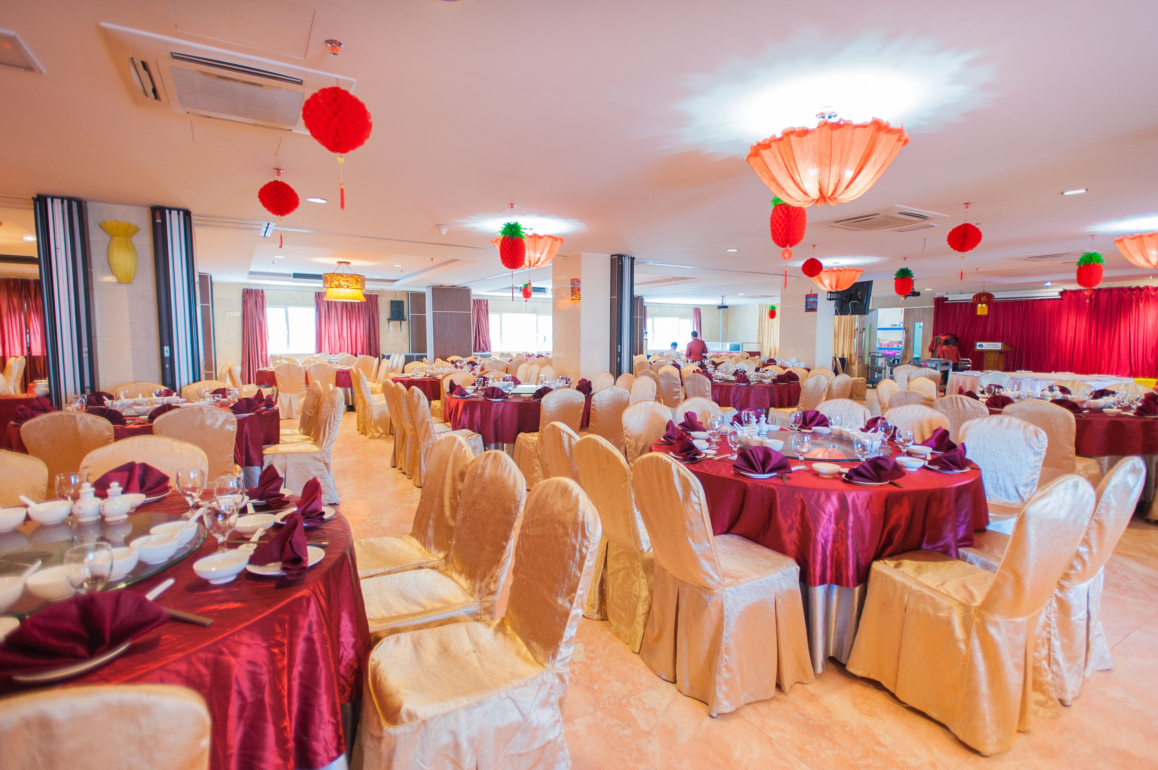 A banquet hall with round tables, chairs, and red decorations.