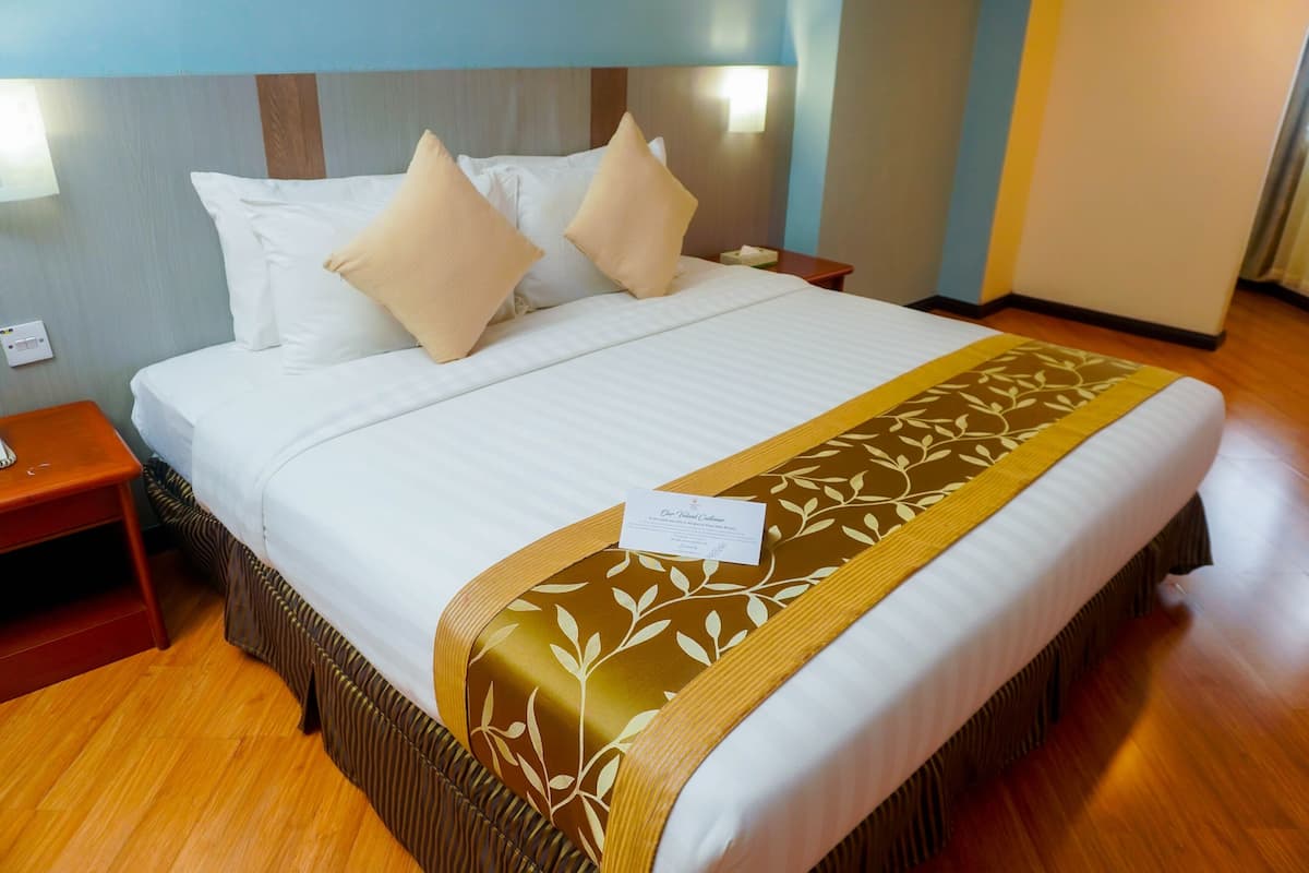 A neatly made bed with a white comforter and two beige pillows, a brown and gold patterned throw blanket, and a small bedside table with a lamp.