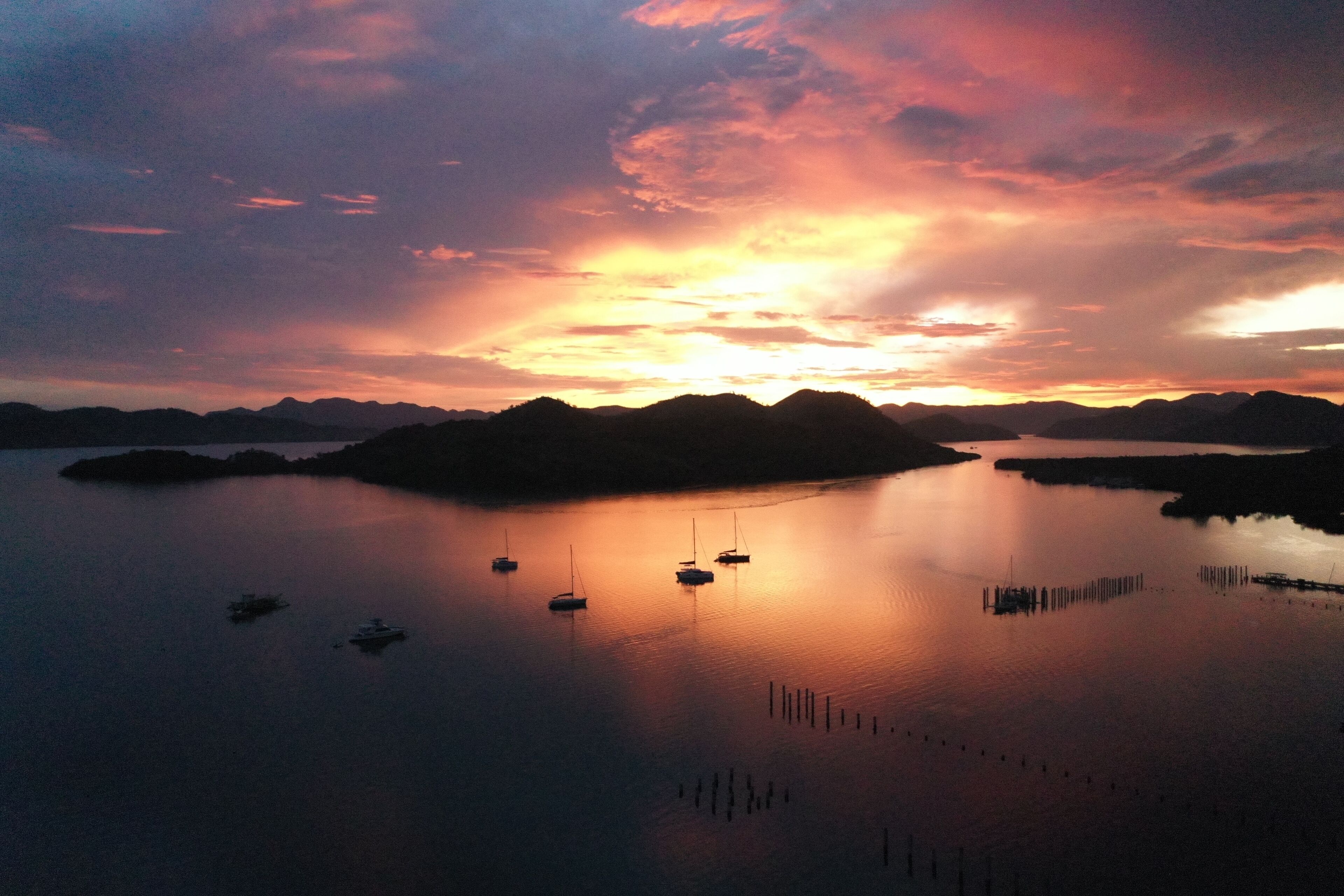 A calm lake with several boats anchored, surrounded by hills and a sunset sky.