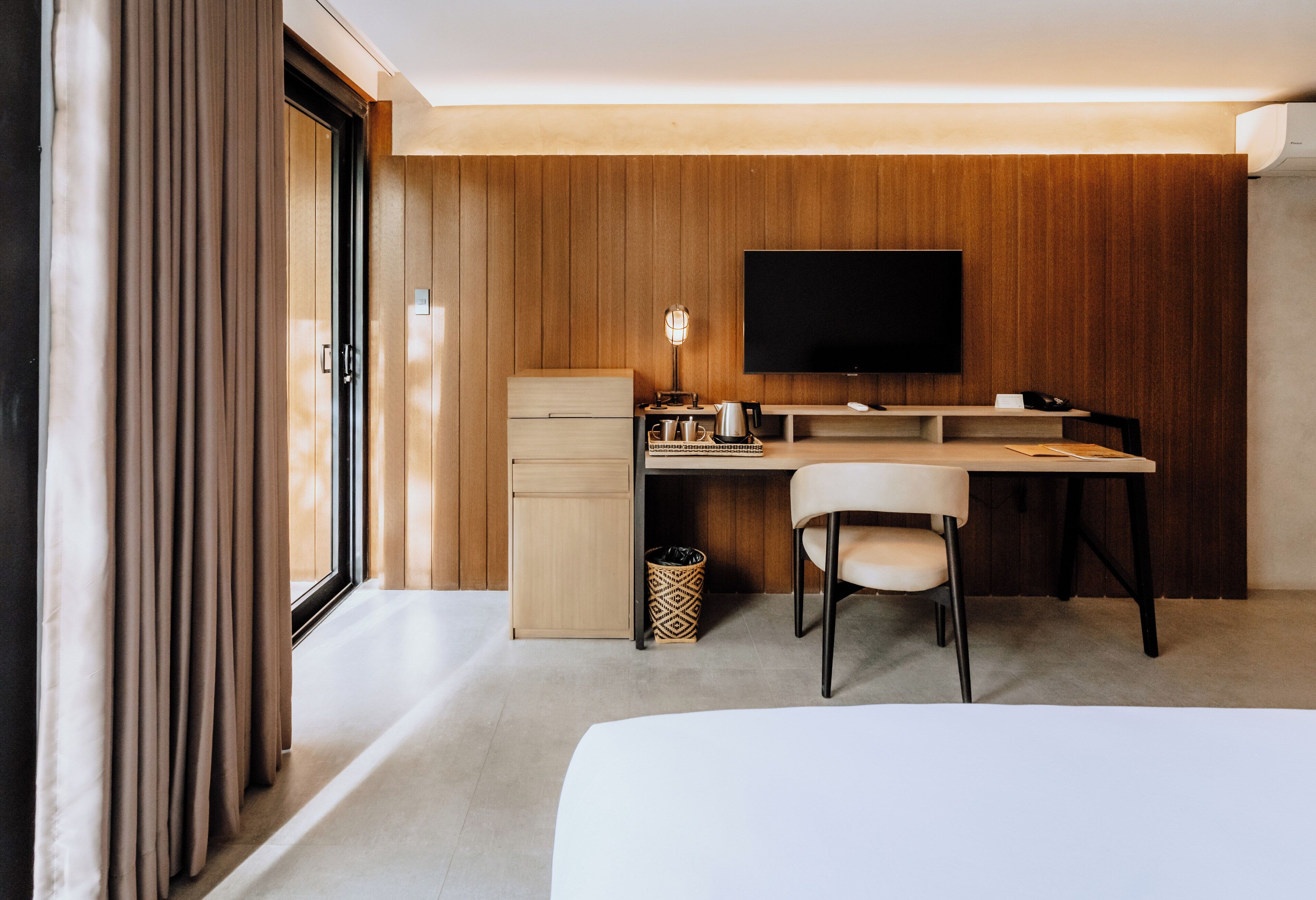 A modern hotel room with a wooden panel wall, a flat-screen TV mounted above a desk, a chair, and a bed.