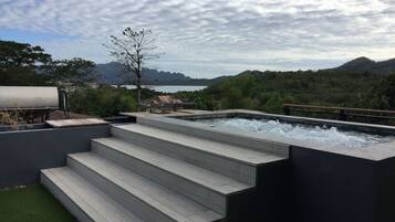 A hot tub with steps leading to it, surrounded by a pool and a view of mountains and a lake.