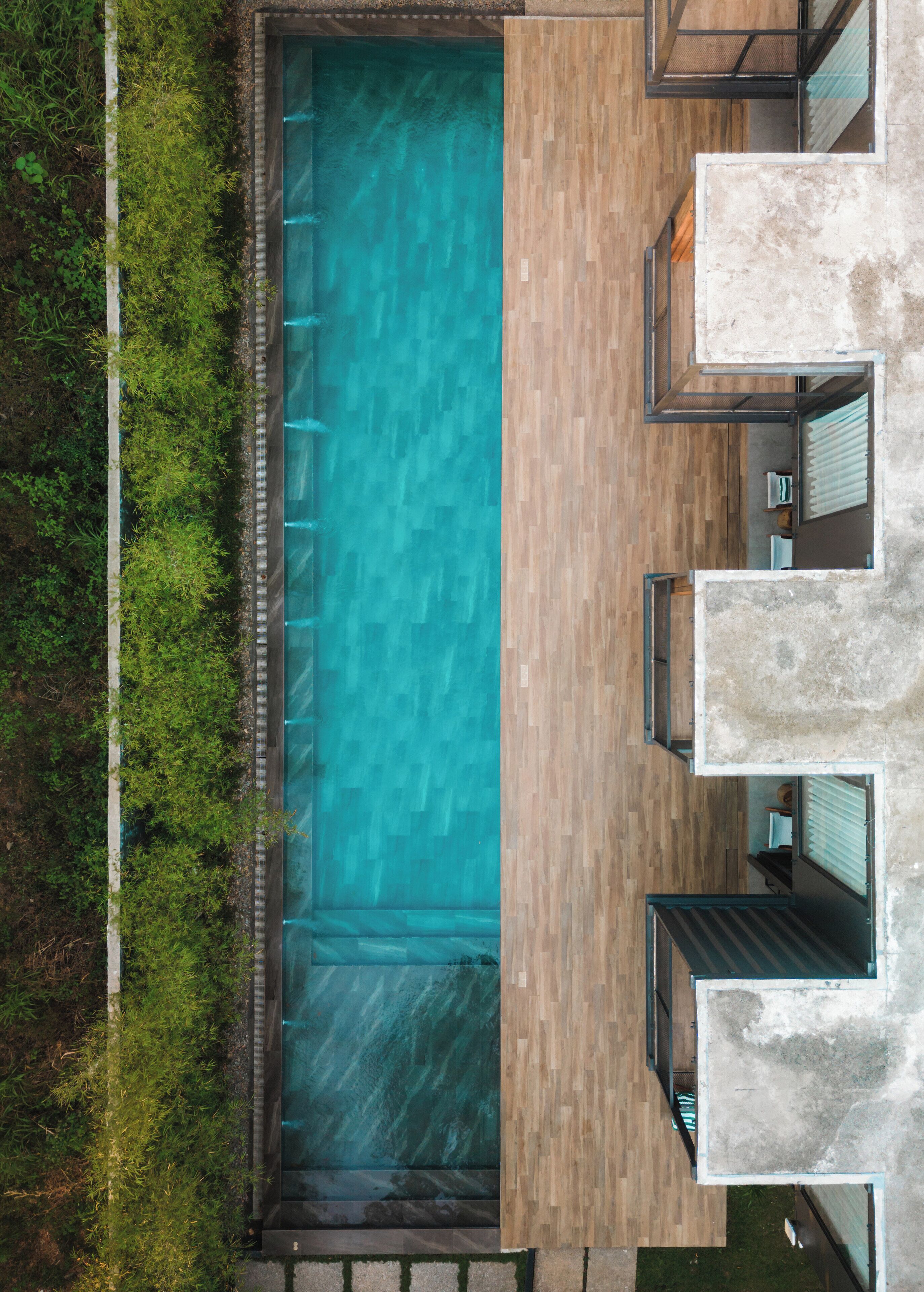 An aerial view of a modern outdoor swimming pool with a wooden deck, surrounded by greenery.
