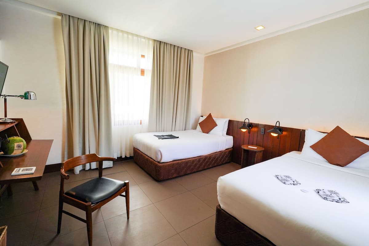 A hotel room with two beds, a wooden chair, a desk with a lamp, and a window with curtains.