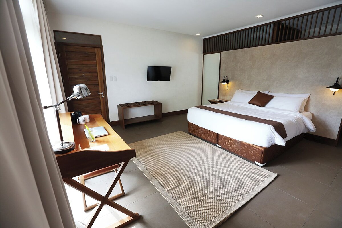 Une chambre d’hôtel avec un lit, un bureau doté d’une lampe, une petite table et un téléviseur fixé au mur.