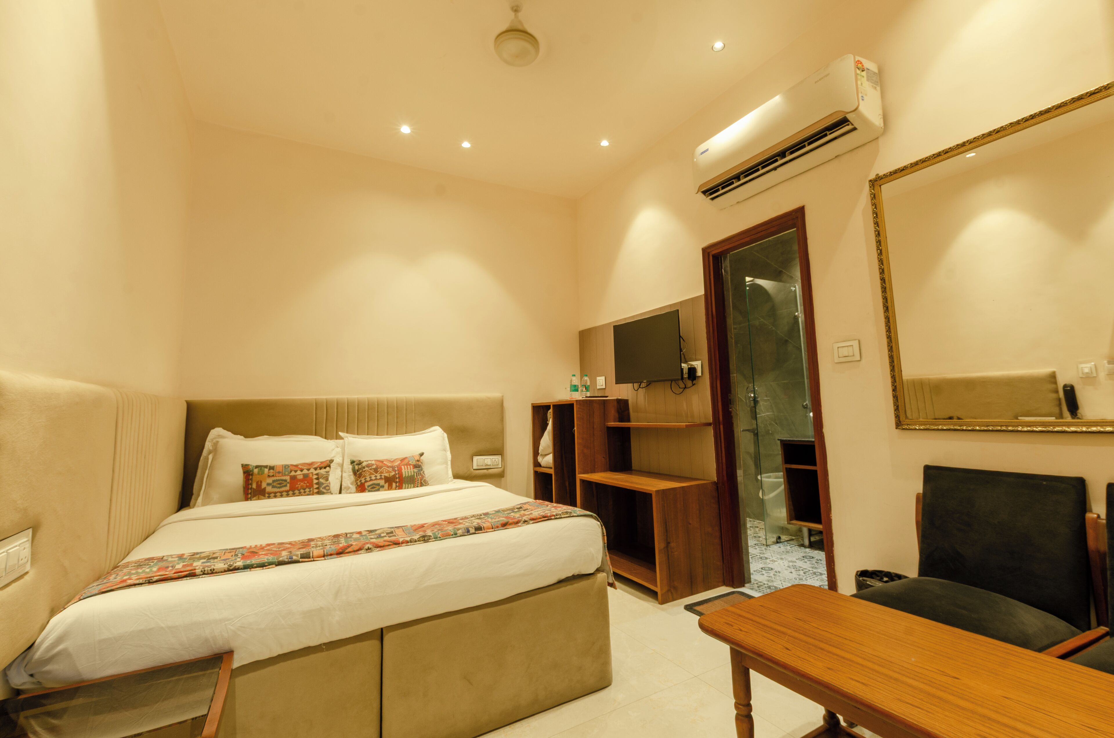 A hotel room with a bed, a desk, a chair, a TV, a mirror, and an air conditioning unit.