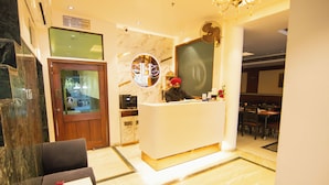 A hotel reception area with a marble reception desk, a receptionist, and a dining area in the background. - Jullundur Hotel (Chandigarh)