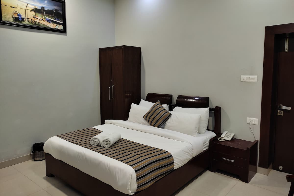 A neatly made bed with white and striped bedding, a wooden wardrobe, a bedside table with a phone, and a painting on the wall.