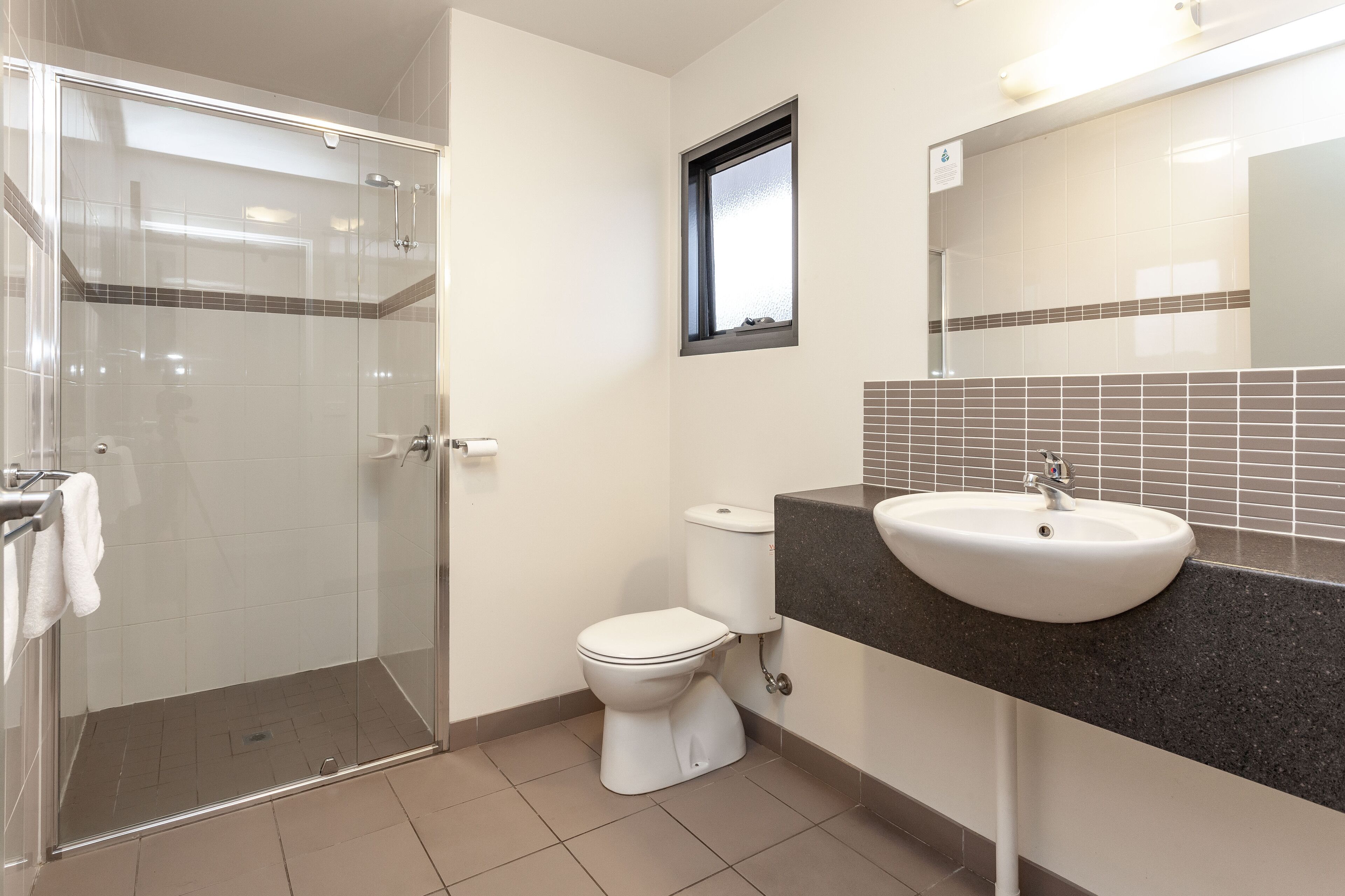 Un baño con ducha separada por mampara de vidrio, inodoro blanco y lavamanos con encimera oscura.