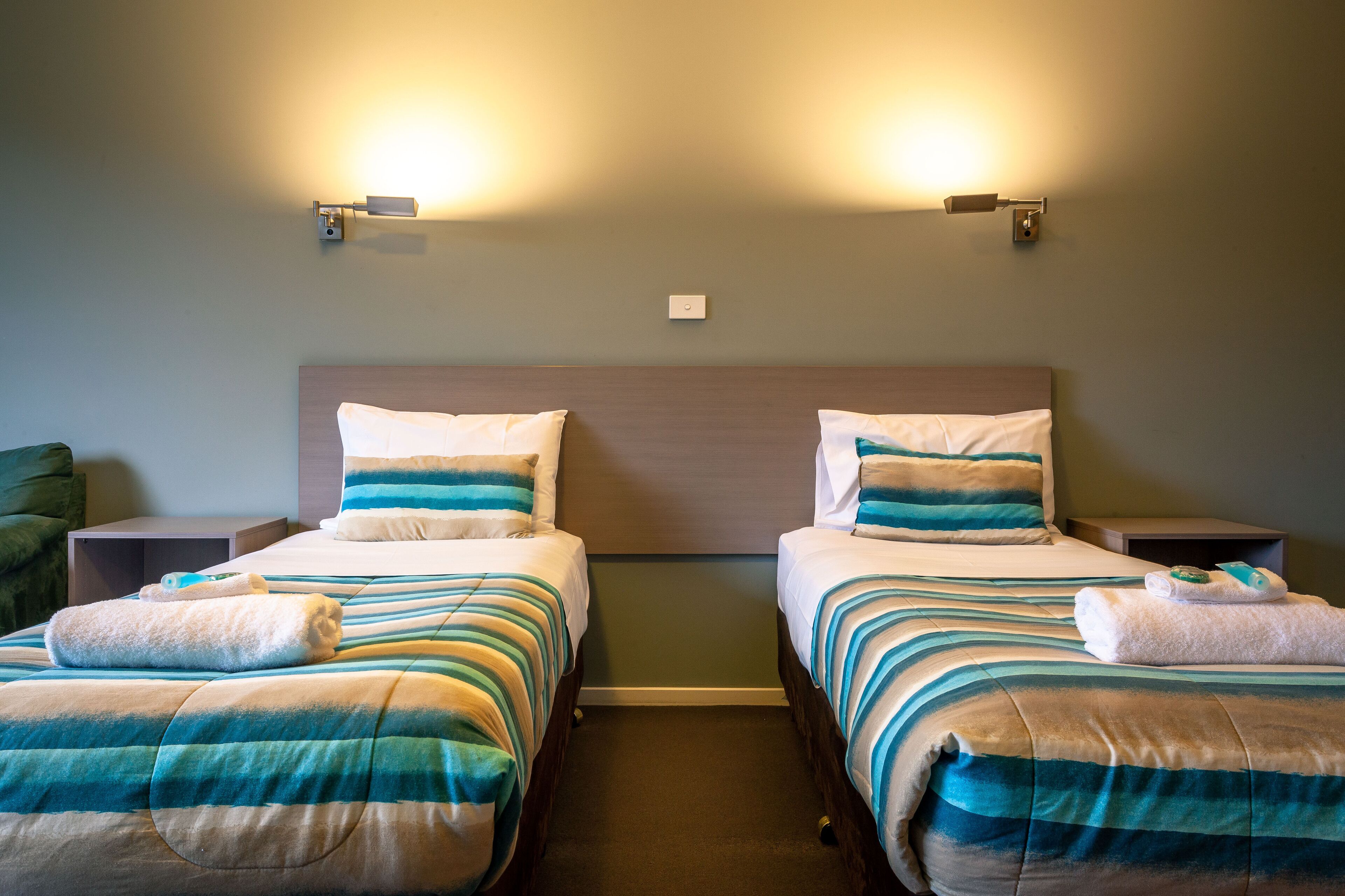 A hotel room with two beds, each with striped bedding and pillows, bedside tables, and wall-mounted lighting.