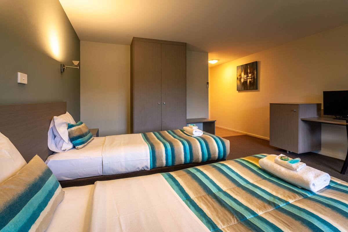 Quarto de hotel com duas camas, um guarda-roupa, uma mesa de trabalho com televisão e um quadro na parede.