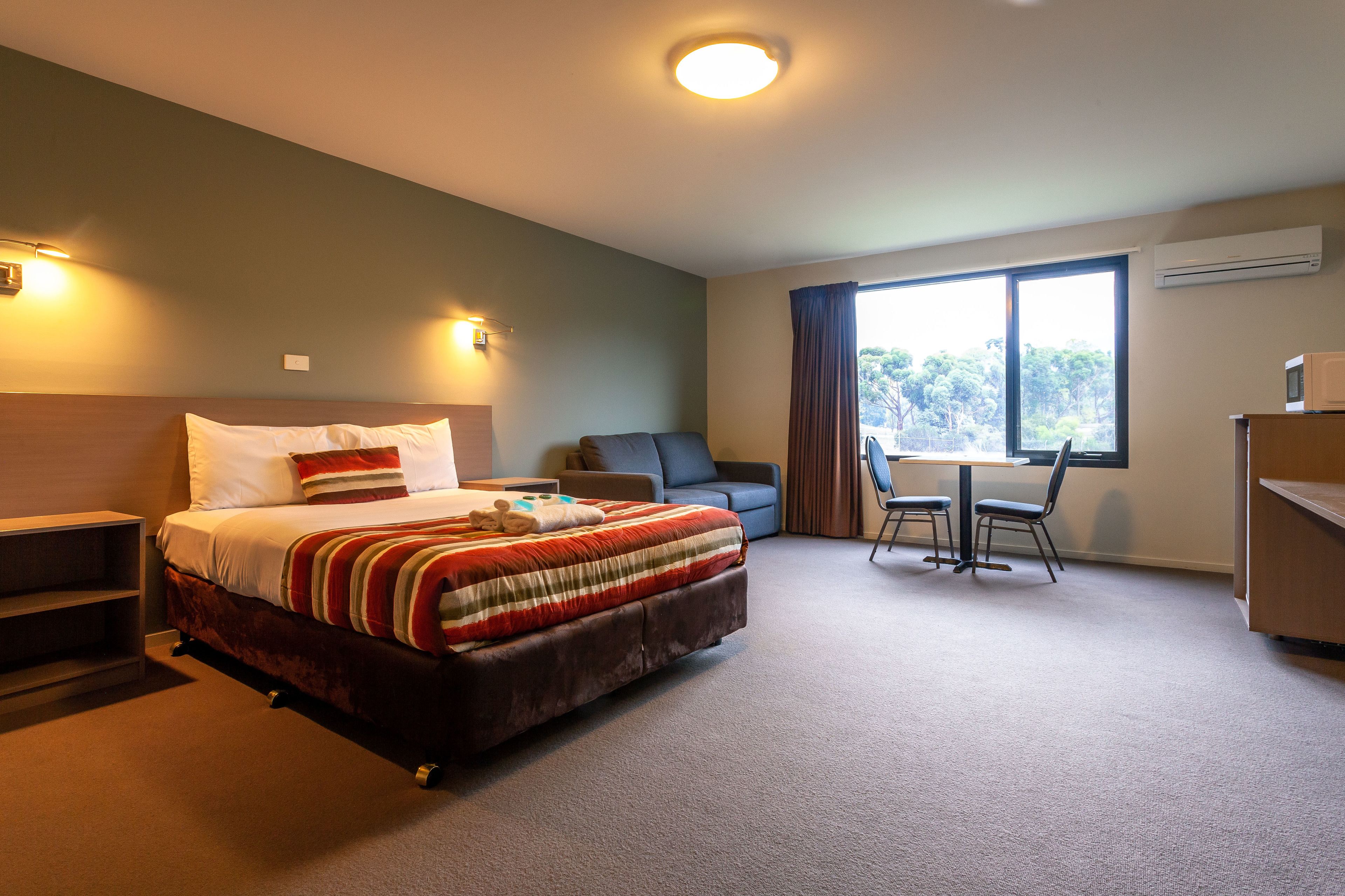 Habitación de hotel con cama, escritorio, silla, sofá y ventana con vistas a árboles.