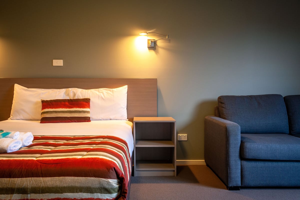 Quarto de hotel com cama, mesa de cabeceira, poltrona azul e luminária fixada na parede.