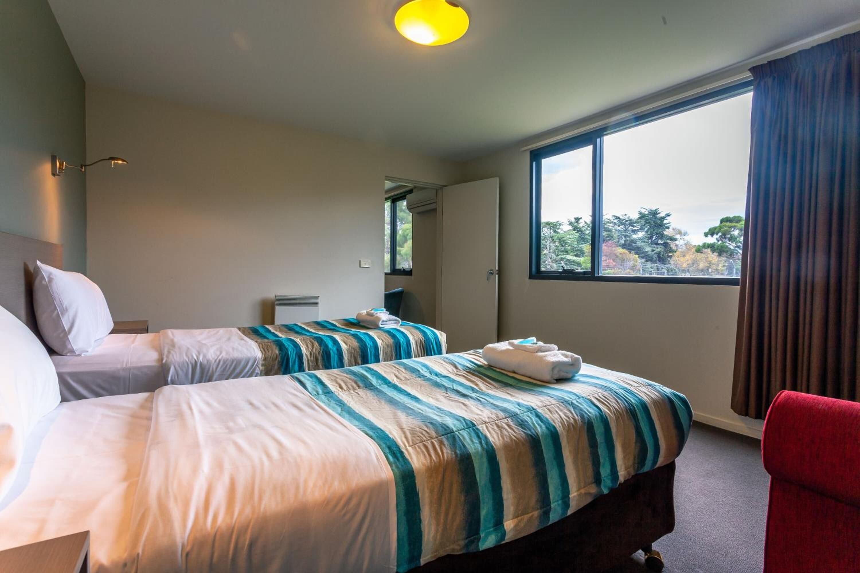 A hotel room with two beds, a red chair, and a large window with a view of trees.