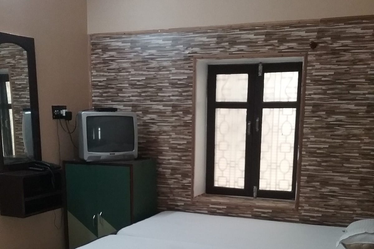 A bedroom with a bed, a television on a cabinet, a window with patterned curtains, and a mirror on the wall.