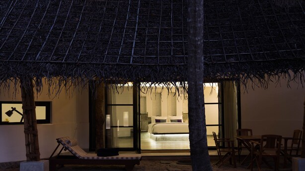 Een kamer aan het strand met rieten dak, een bed, een lounge stoel en een eethoek.