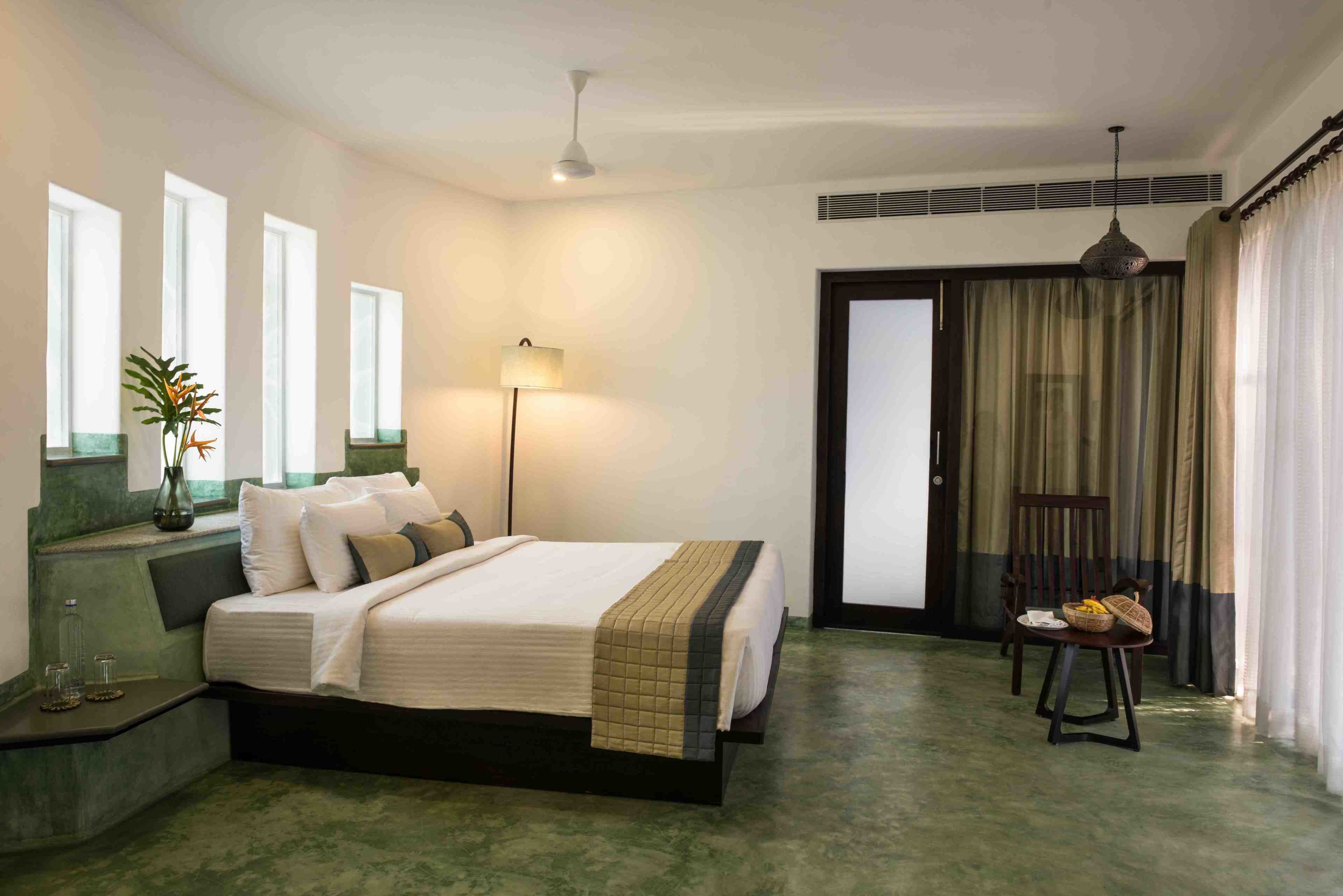 A bedroom with a bed, bedside tables, a floor lamp, a chair, and a view of the outside through a door.