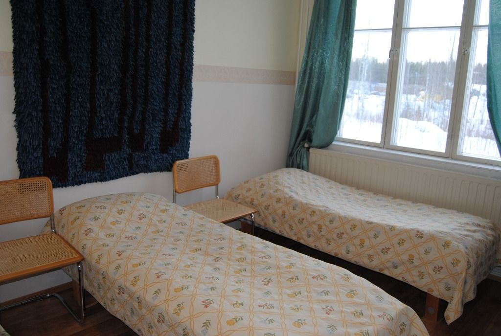 A room with two single beds, a chair, a window with curtains, and a radiator.