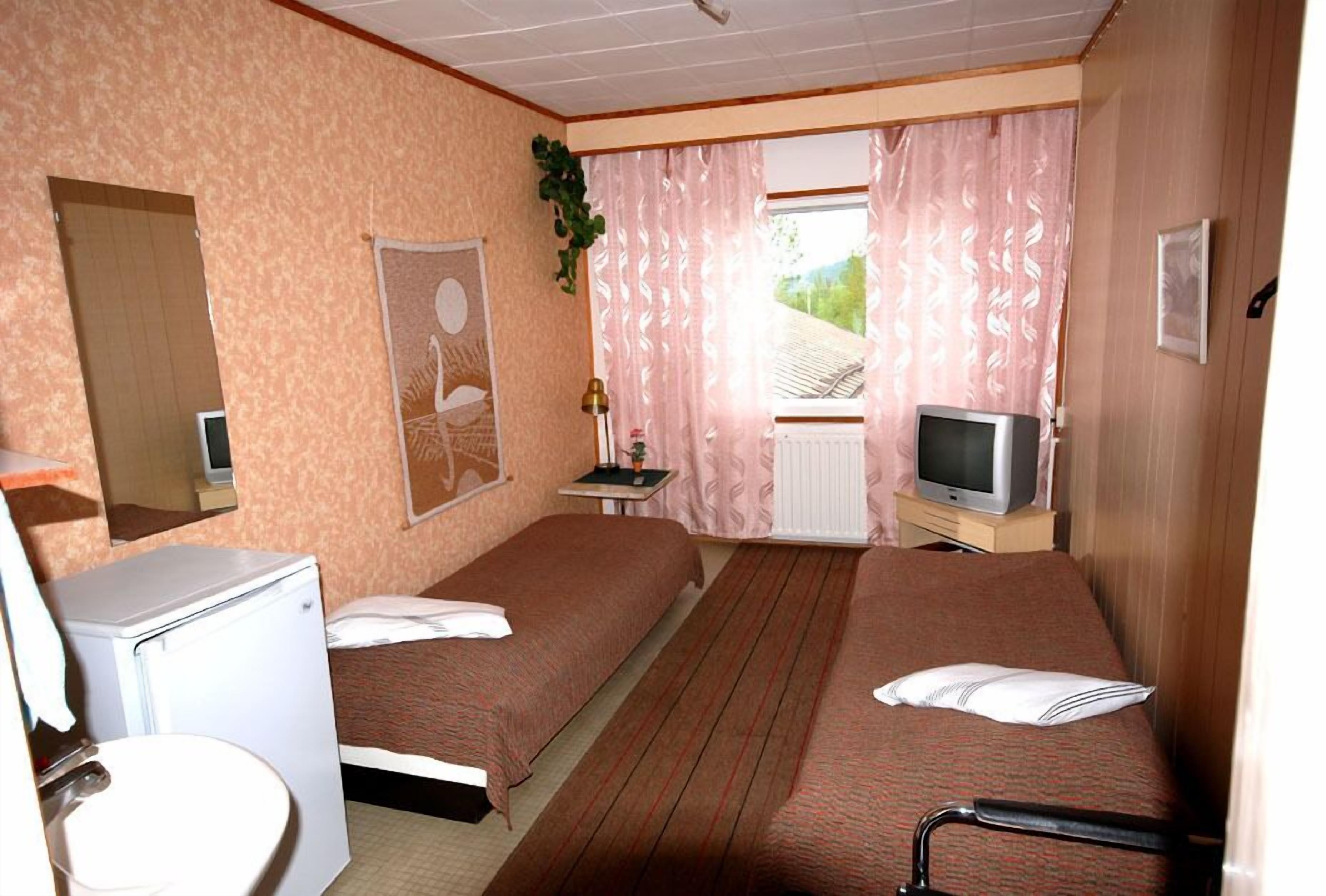 Habitación de hotel con dos camas, una mesita con una lámpara, un televisor y una ventana con cortinas.