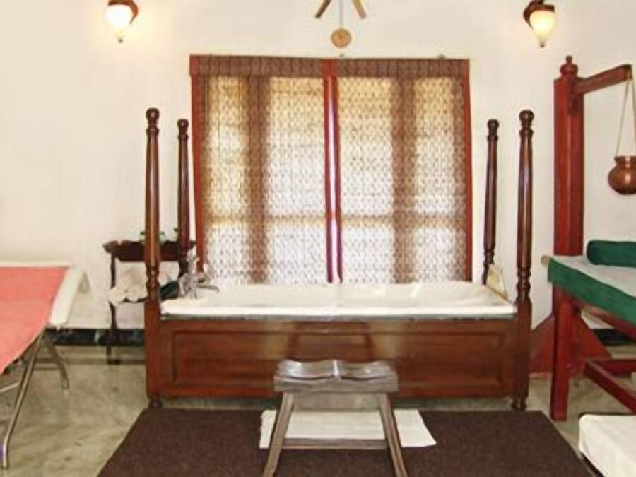 A room with a wooden bed, a small table, a chair, a window with curtains, and a wall-mounted shelf.