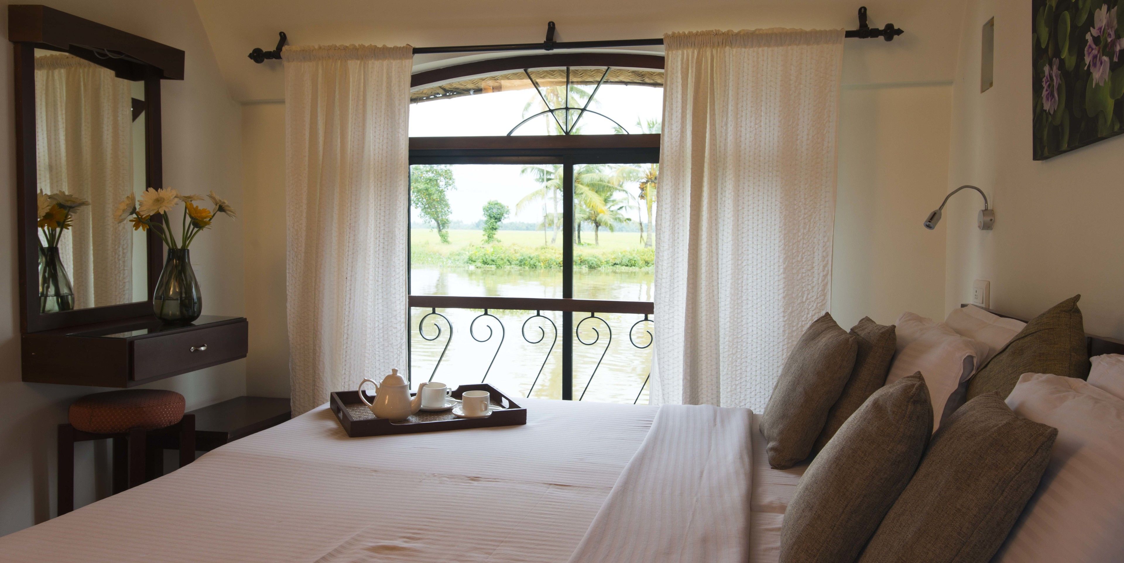 Une chambre d’hôtel avec un lit, une table de chevet sur laquelle se trouve un plateau avec du thé et des tasses, un miroir et une fenêtre avec des rideaux.