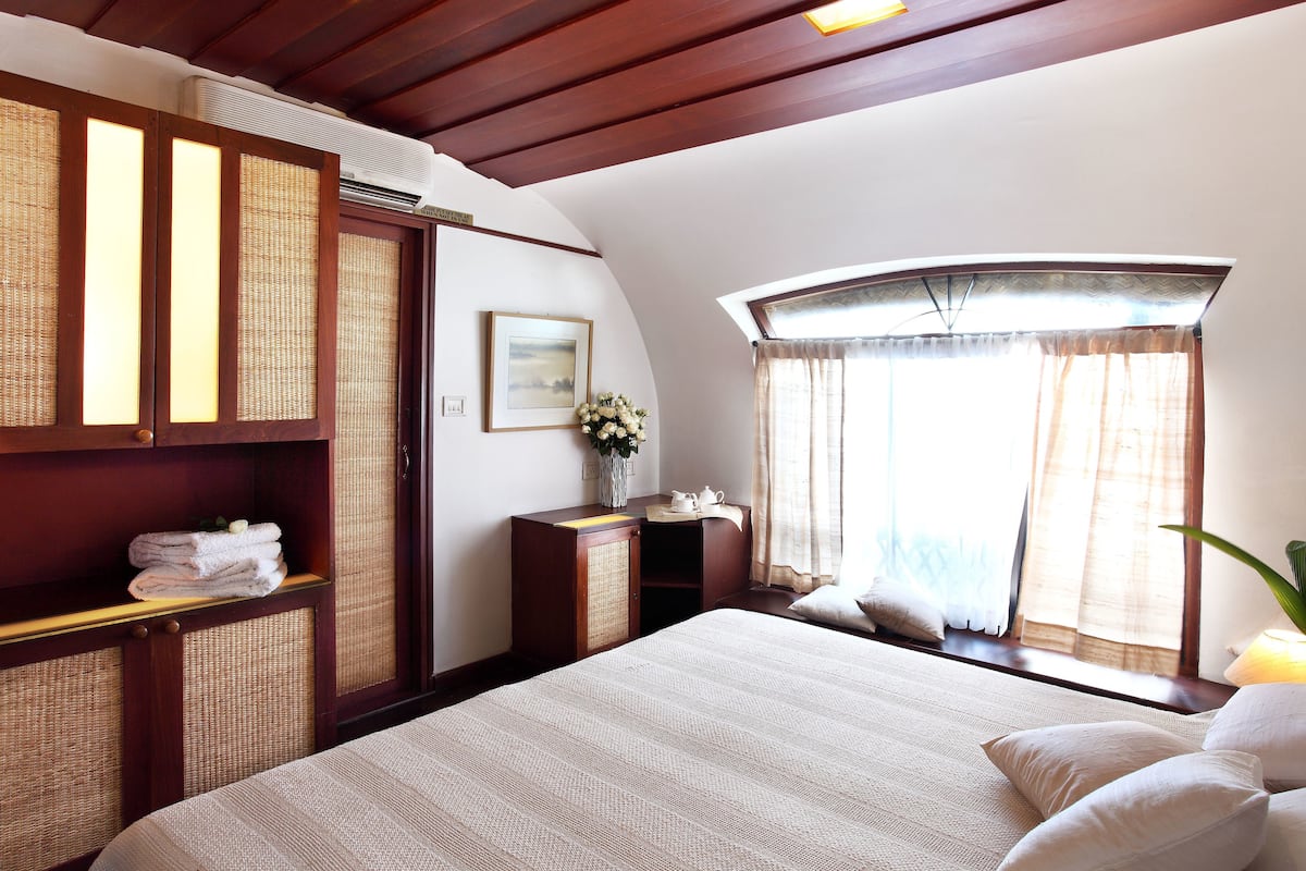 Un dormitorio con una cama grande, un armario de madera, una mesita con un jarrón de flores y una ventana con cortinas.