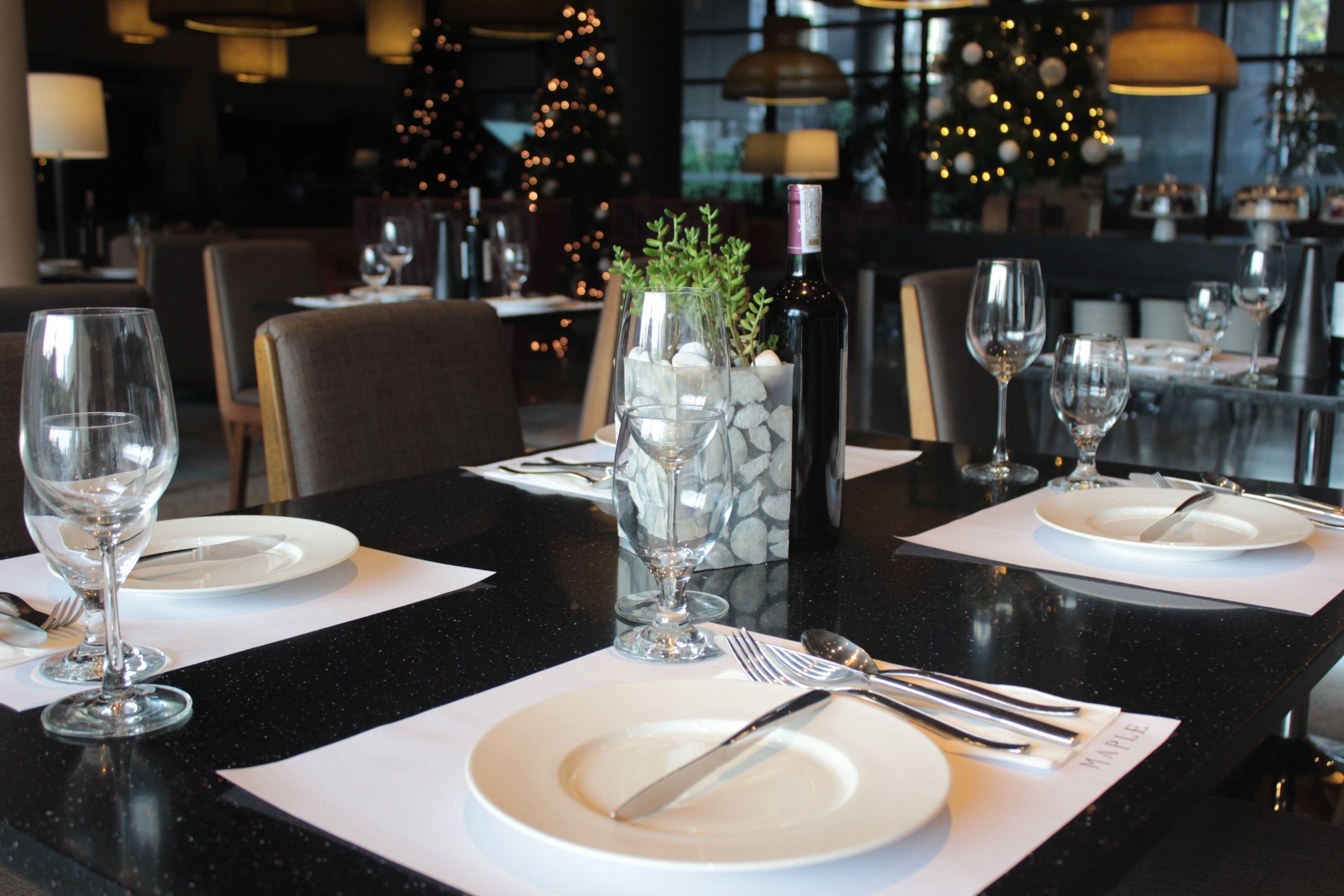 A restaurant table set with white plates, cutlery, and wine glasses, with a decorative vase and a bottle of wine.