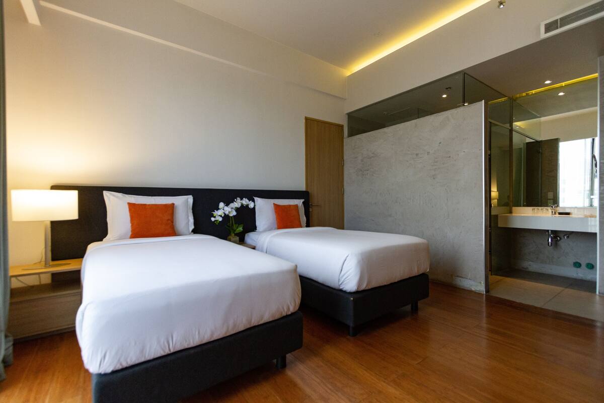 Une chambre d’hôtel moderne avec deux lits, une table de chevet, une lampe et une salle de bain dotée d’une douche à parois de verre.