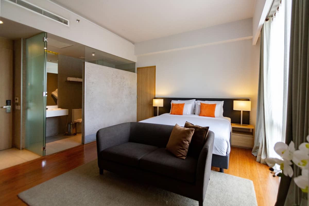 Une chambre d’hôtel moderne comprenant un lit, un canapé, une salle de bain fermée par des parois de verre et une table de chevet avec une lampe.