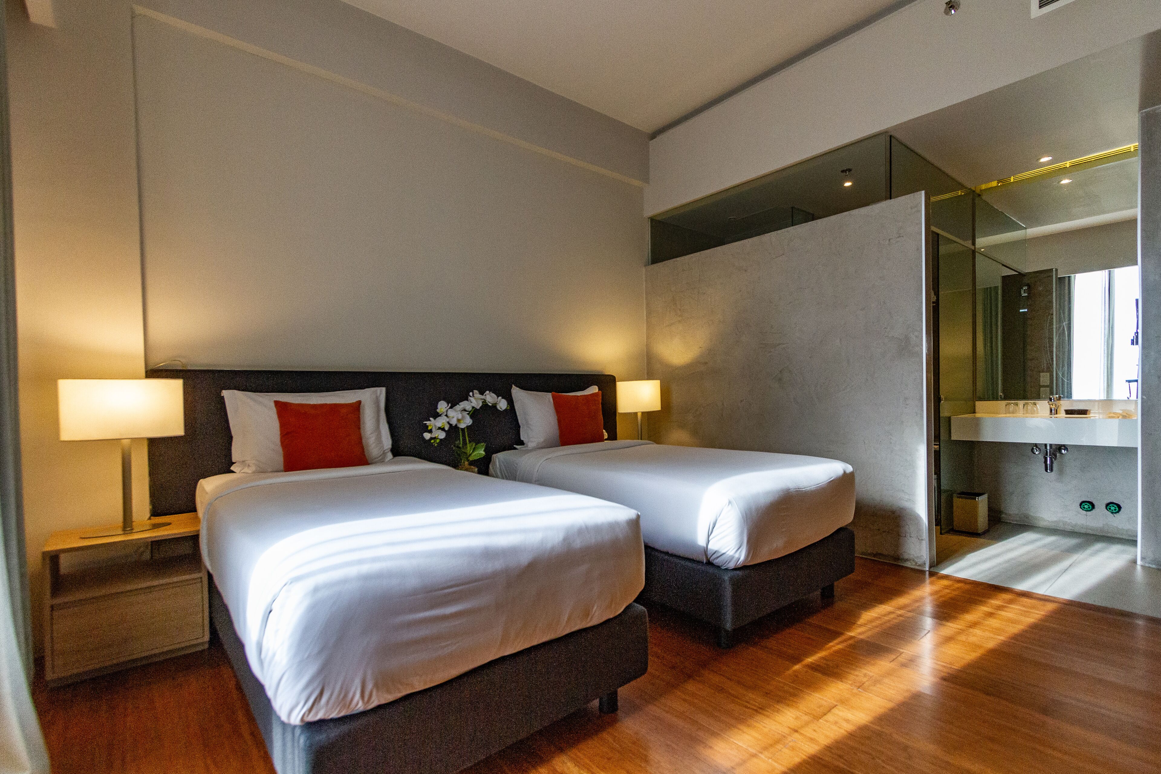 Habitación de hotel con dos camas, una mesita de noche, una lámpara, un espejo y un baño visible a través de una puerta de cristal.