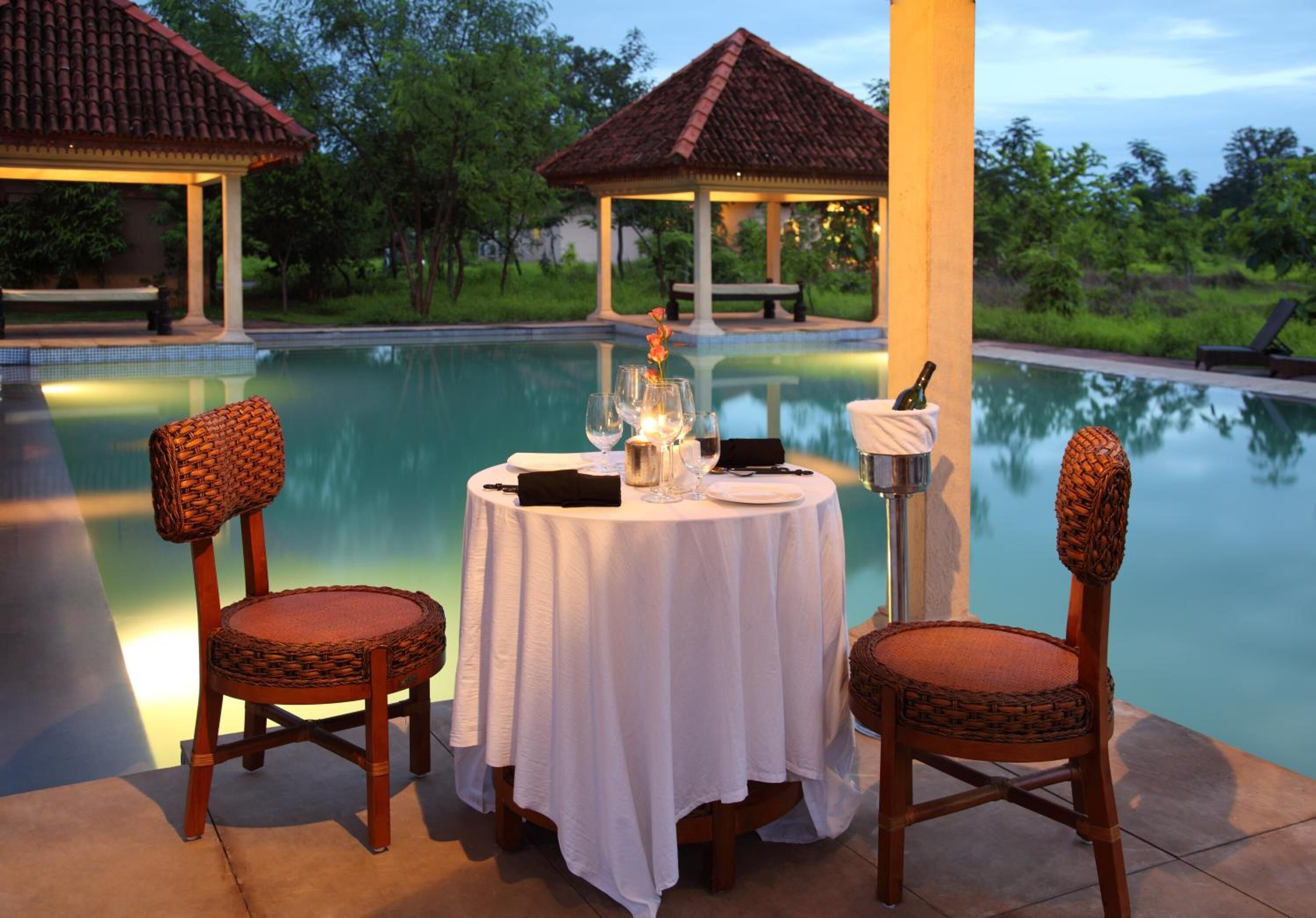 A dining table by a pool with two wicker chairs, a bottle, glasses, and a candle.