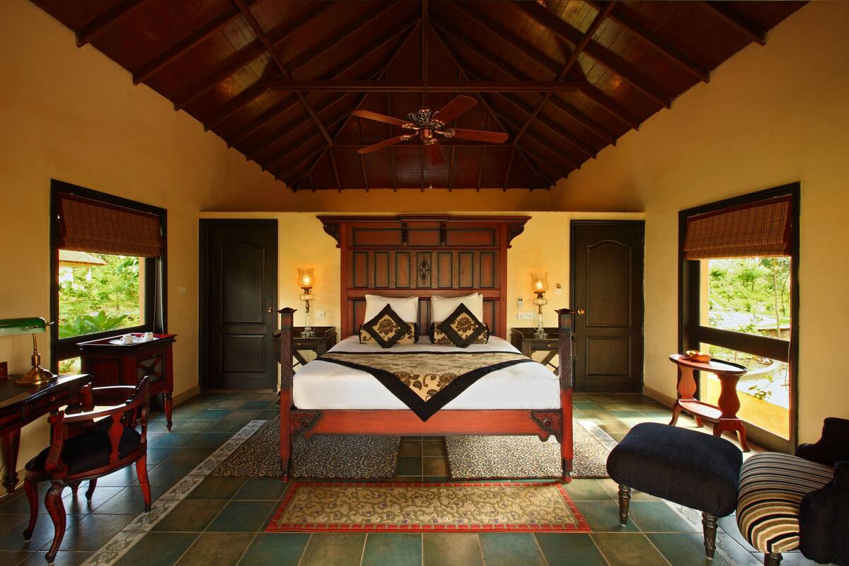 A spacious bedroom with a large bed, wooden headboard, and a ceiling fan. There are two bedside tables with lamps, a desk, and a chair. The room has large windows with views of greenery outside.