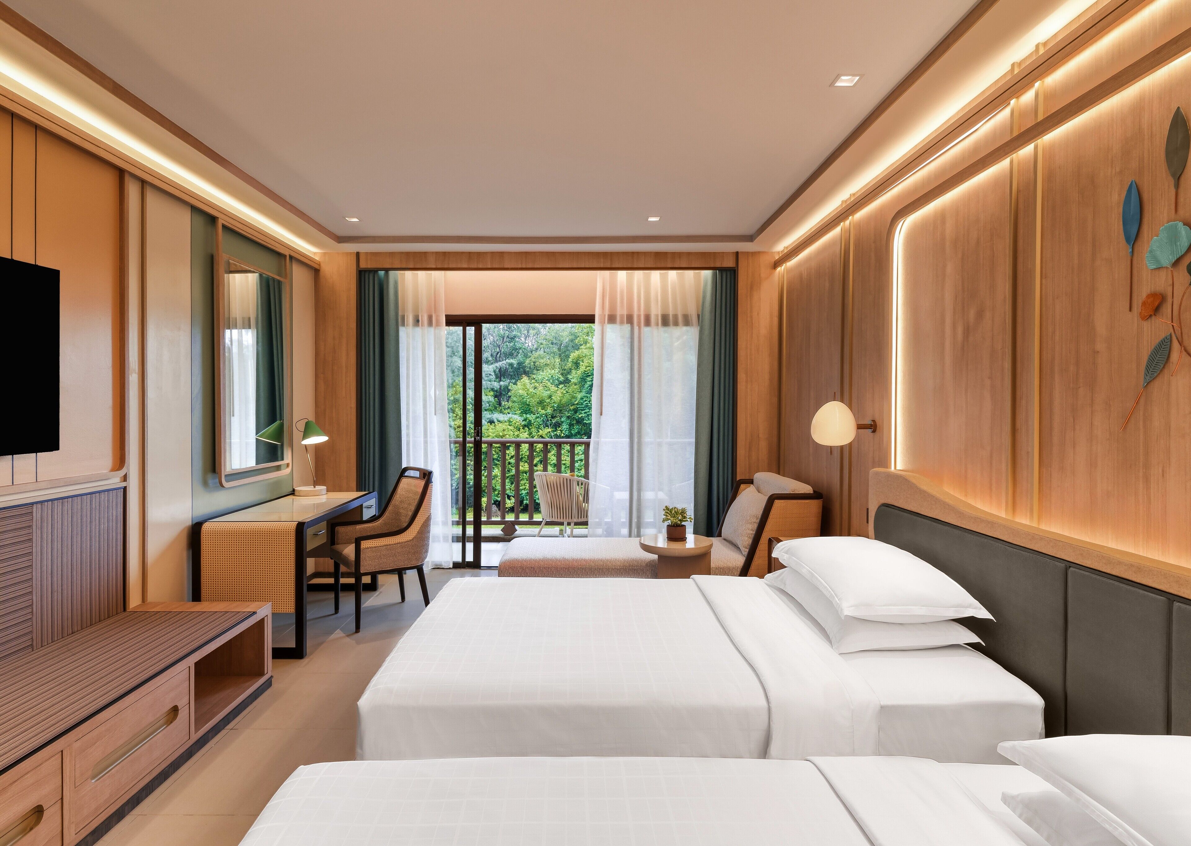 A hotel room with a bed, a desk, a chair, a TV, and a balcony with a view of greenery.