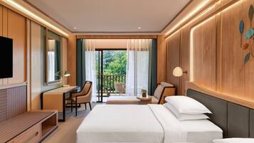 A hotel room with a bed, a desk, a chair, a TV, and a balcony with a view of greenery.