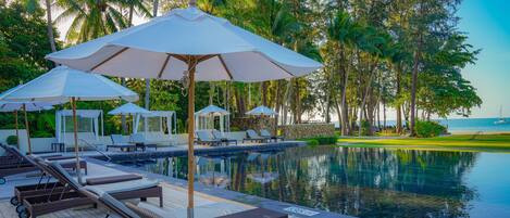 Zona junto a la piscina con sillas de descanso y sombrillas, rodeada de palmeras y vista al mar.