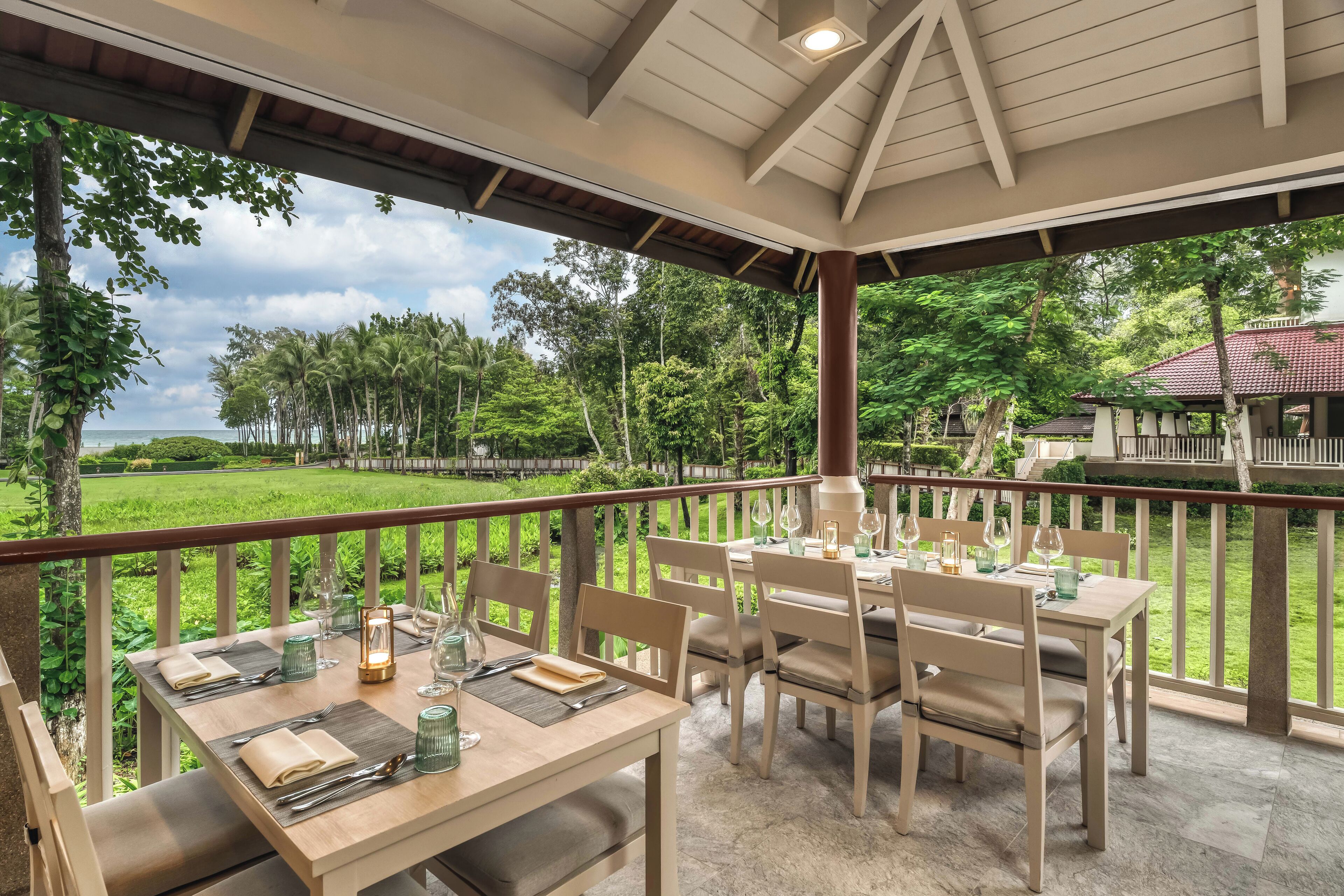A covered outdoor dining area with tables set for two, overlooking a lush garden and a distant building.