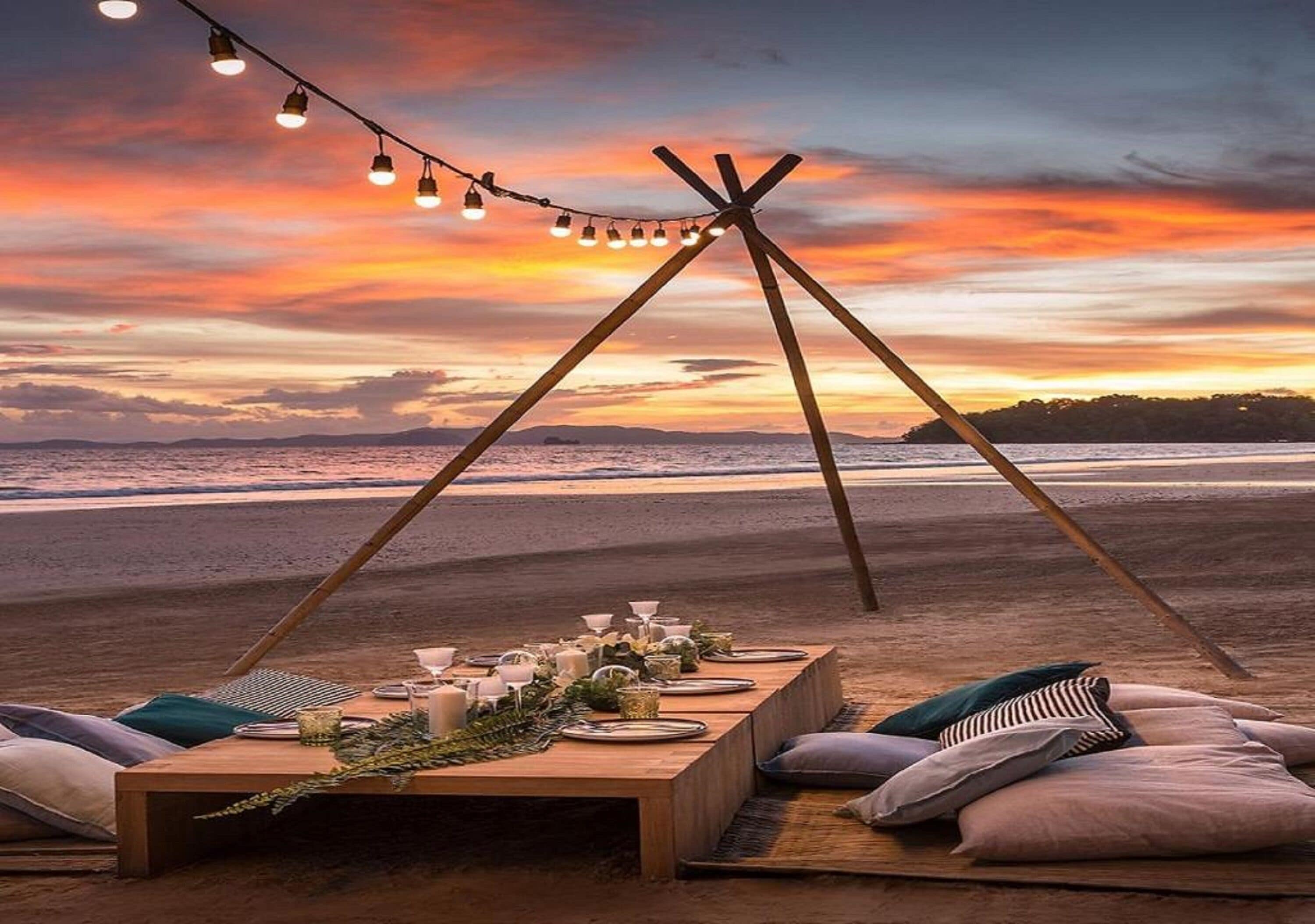 Montaje para comer en la playa con una mesa de madera, luces colgantes y vista al océano al atardecer.