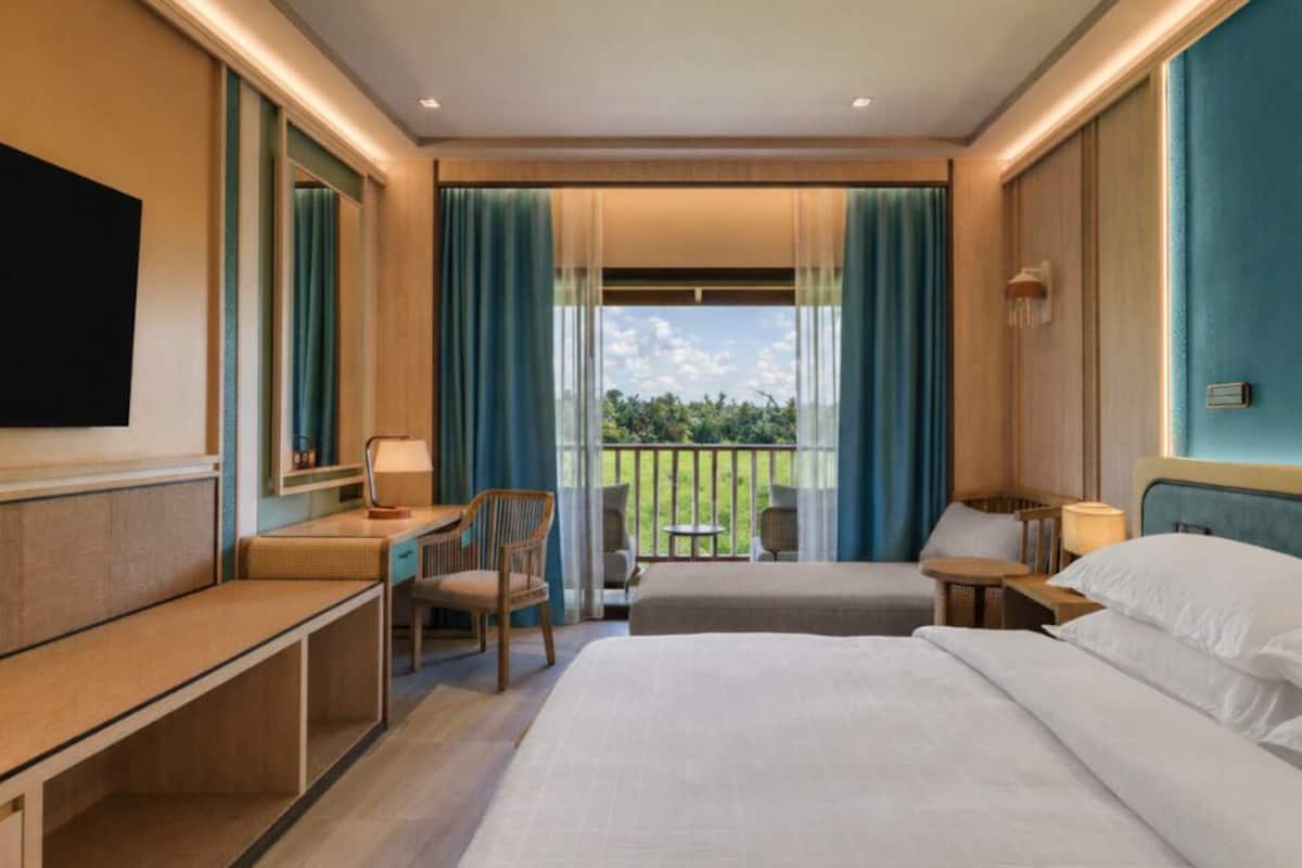 Een hotelkamer met een bed, een bureau, een stoel, een klein tafeltje en een balkon met uitzicht op groen.