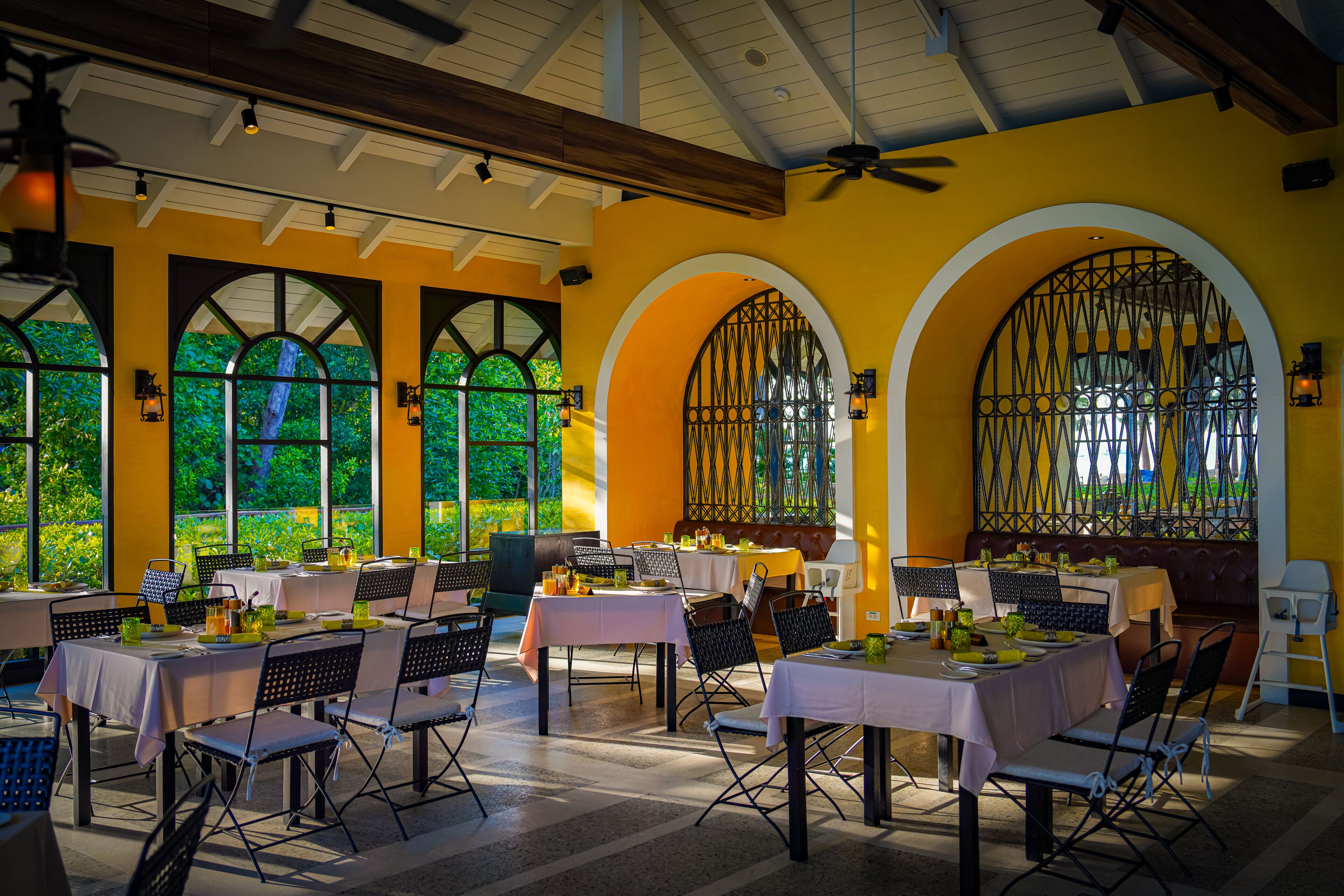 A restaurant with tables set for dining, large windows with greenery outside, and a ceiling fan.