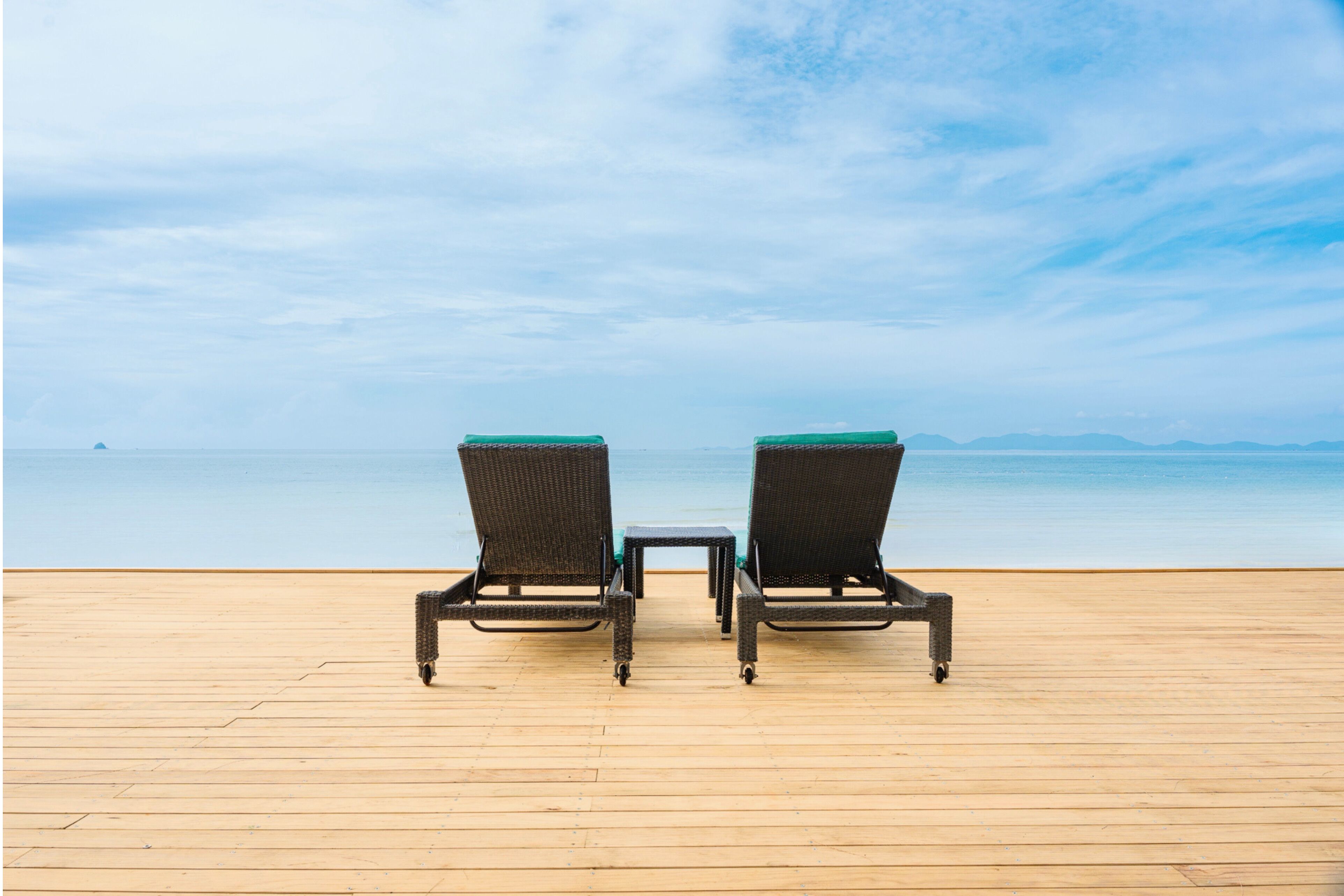 Dos sillones de mimbre mirando a una terraza de madera con vista al mar y montañas distantes.