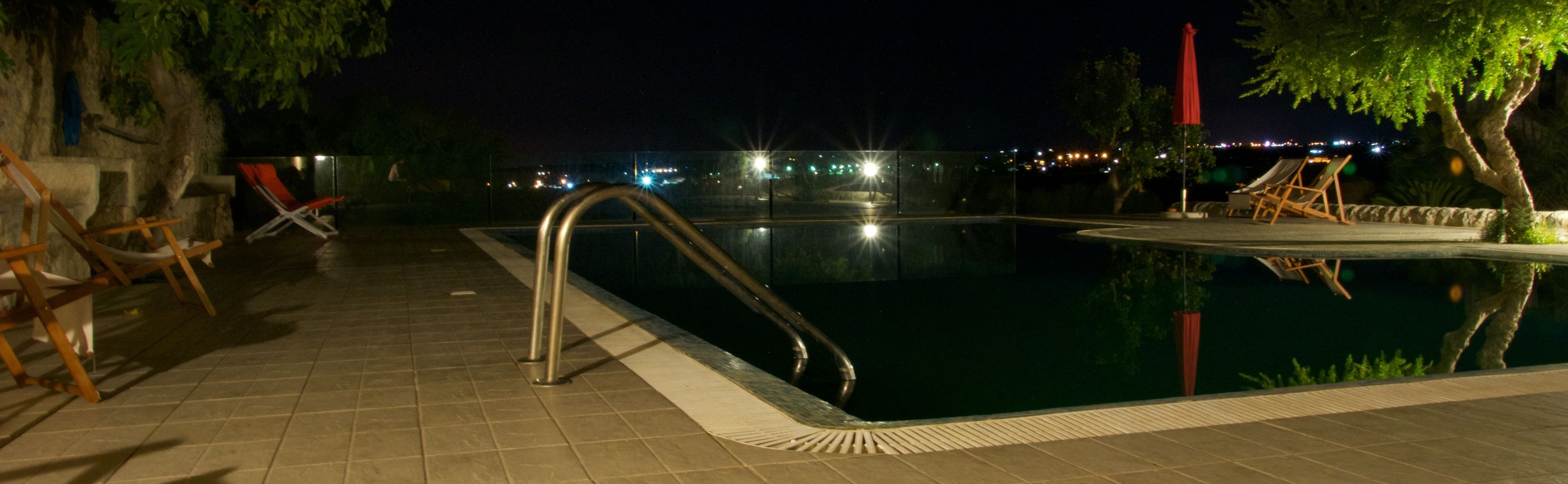 Un espace piscine nocturne avec un escalier en métal, des chaises longues et une vue sur les lumières de la ville en arrière-plan.