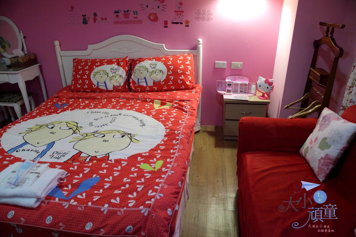 A bedroom with a bed covered in a red heart-patterned bedspread, a white headboard, and a pink wall with various decorations.