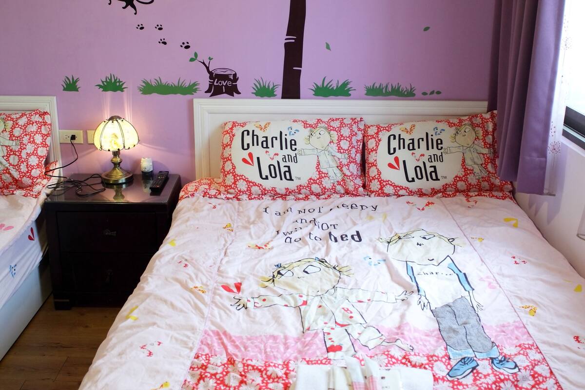 A bedroom with a bed, two pillows, a nightstand, a lamp, and wall decorations including a tree, leaves, and a cat.