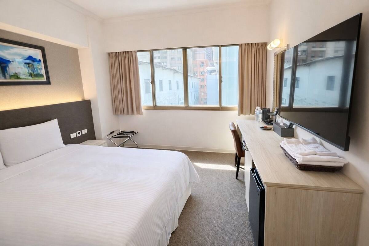 A hotel room with a bed, a desk, a chair, a mirror, and a window with curtains.