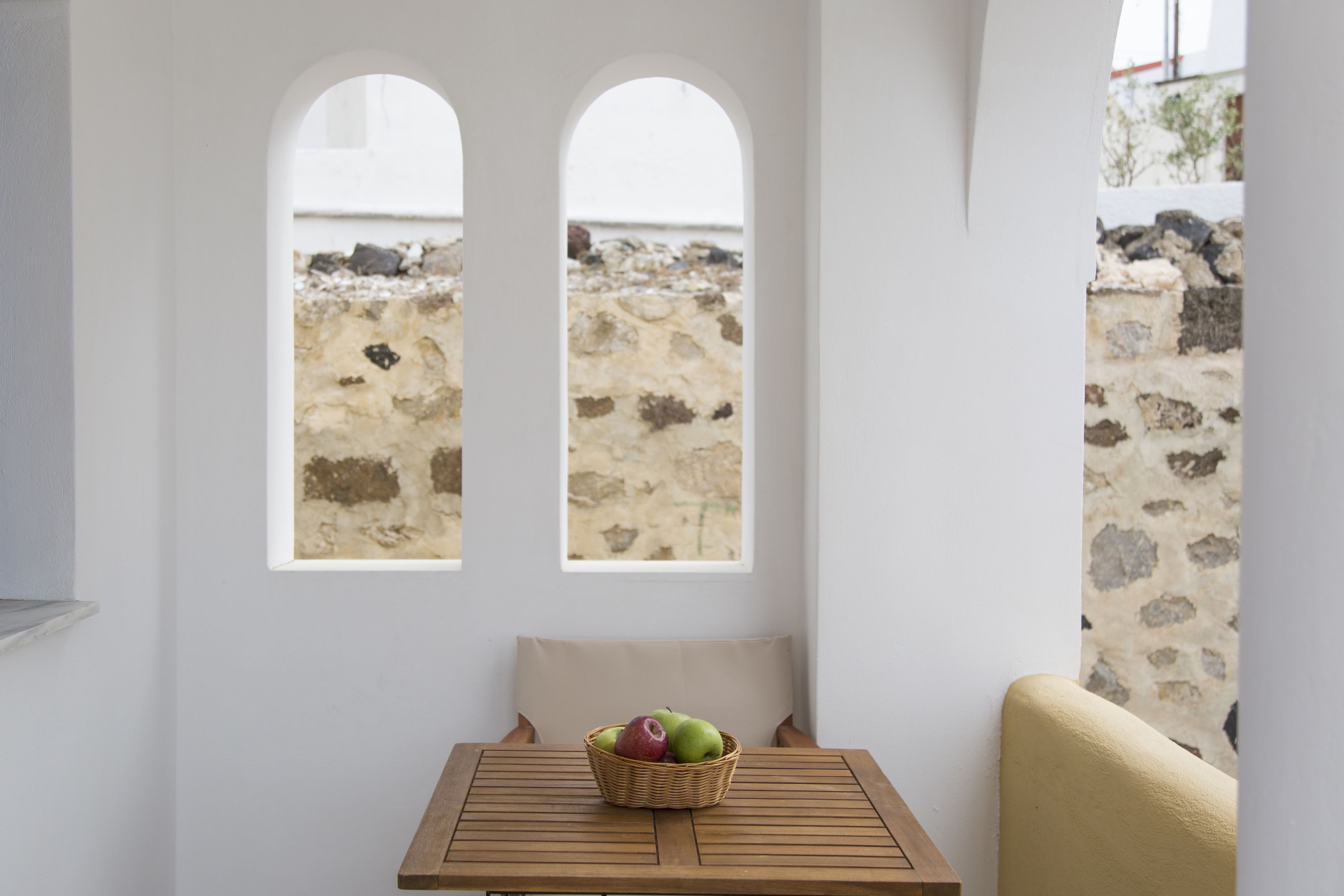 Ein Zimmer mit einem Holztisch, einem Korbwesen mit Äpfeln und Blick durch Bogenfenster auf eine Steinmauer.
