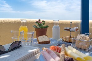 Una mesa con alimentos para el desayuno, como pan, tomates y queso, junto con una planta en maceta y vasos de jugo.