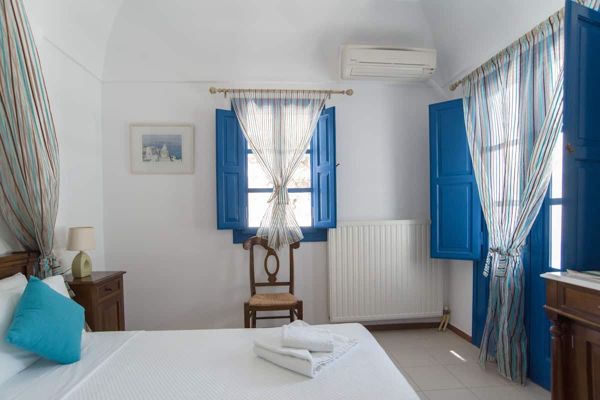 A hotel room with a bed, a chair, a nightstand, a picture on the wall, and a window with blue shutters.