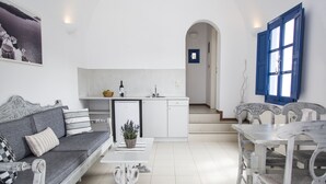 Una sala moderna con un sofá gris, una mesa de centro blanca y una mesa de comedor con sillas. Se ve un área de cocina con armarios blancos y un lavamanos al fondo.