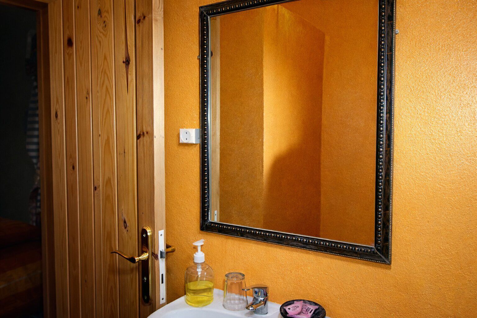 A bathroom with a wooden door, a mirror with a black frame, a sink with a soap dispenser, and a glass pitcher.